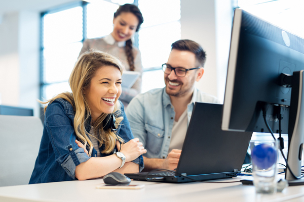 zwei Frauen und ein Mann, die lachend um einen PC-Arbeitsplatz sitzen und stehen
