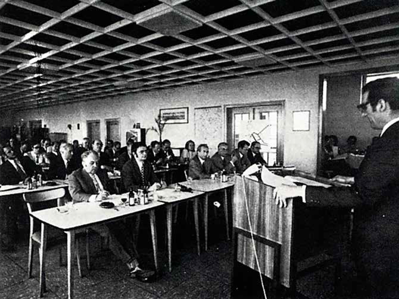 ein altes Schwarzweiß-Foto eines Mannes, der an einem Rednerpult steht und vor einer Gruppe an Tischen sitzenden Männern spricht