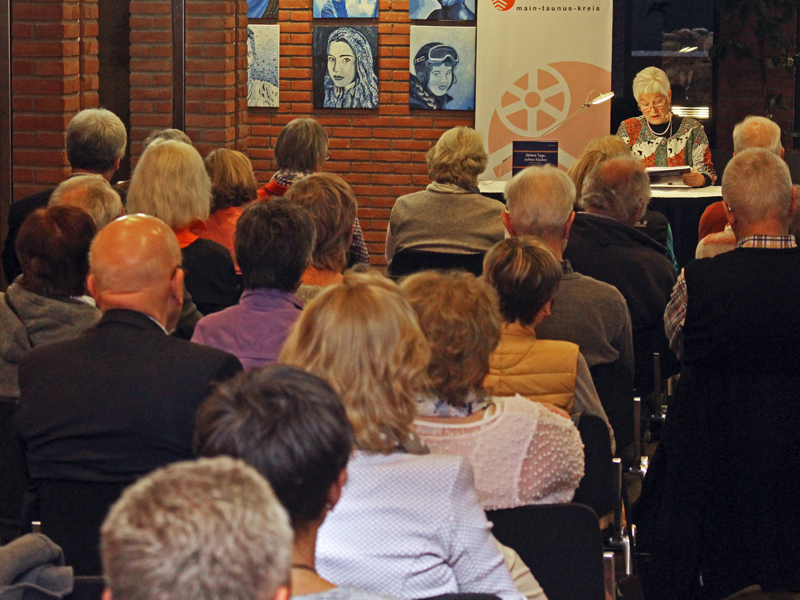 eine grauhaarige Dame, die vor Publikum aus einem Buch liest