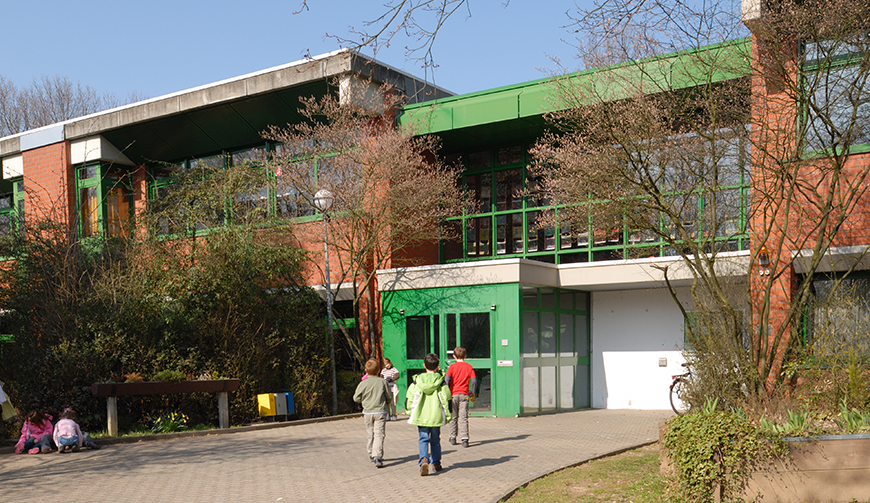 Blick über einen Hof und eine Wiese auf ein Gebäude, das teilweise grün gestrichen ist; im Vordergrund einige Kinder mit dem Rücken zur Kamera