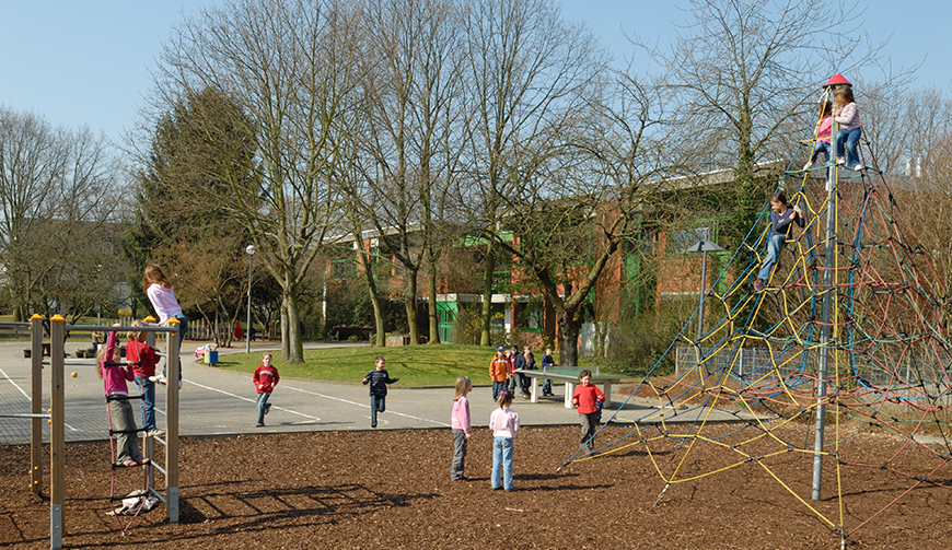 Blick auf eine Fläche mit Spielgeräten und Klettermöglichkeiten, auf der einige Kinder spielen; im Hintergrund Bäume und Gebäude