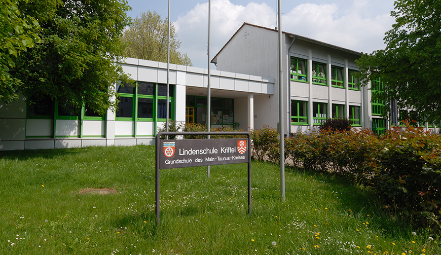 Schild mit der Aufschrift Lindenschule auf einer Wiese; im Hintergrund Fahnenmasten und ein Gebäude