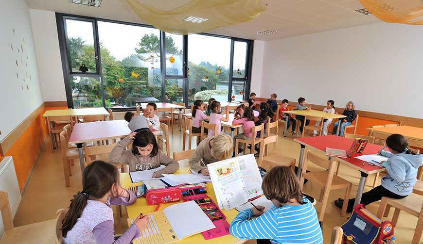 einen Raum mit großen Fenstern, in dem Kinder in Gruppen an Tischen sitzen und arbeiten