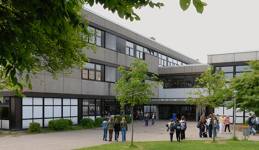 Blick über einen von Bäumen gesäumten Schulhof auf ein graues Gebäude