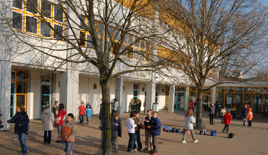 Kinder in einem mit Bäumen bestandenen Innenhof; im Hintergrund ein großes Gebäude