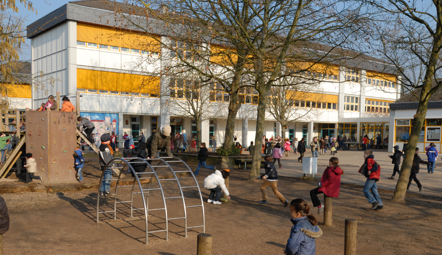 ein Spielplatz mit Klettergerüsten, auf dem Kinder spielen; im Hintergrund ein großes Gebäude
