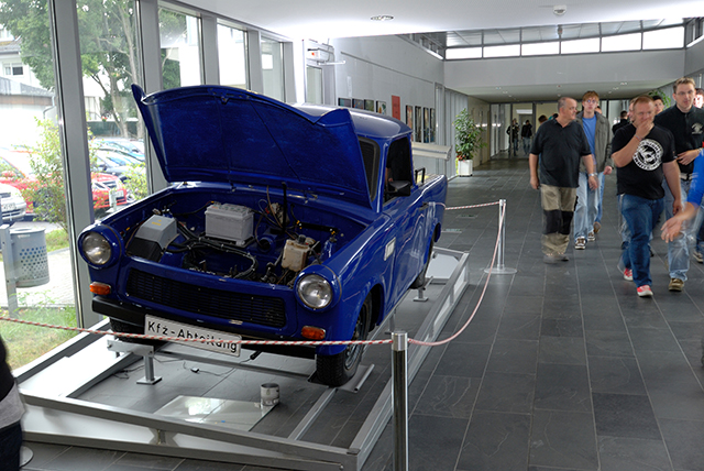 ein älteres blaues Auto mit aufgeklappter Motorhaube in einem großen Ausstellungsraum, im Hintergrund einige Personen