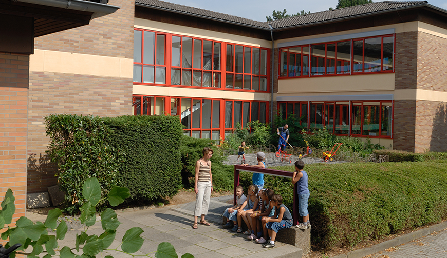 Blick auf einen mit Hecken umwachsenen Schulhof, in dem sich einige Schüler aufhalten; umrahmt wird das Gelände von mehreren Gebäuden