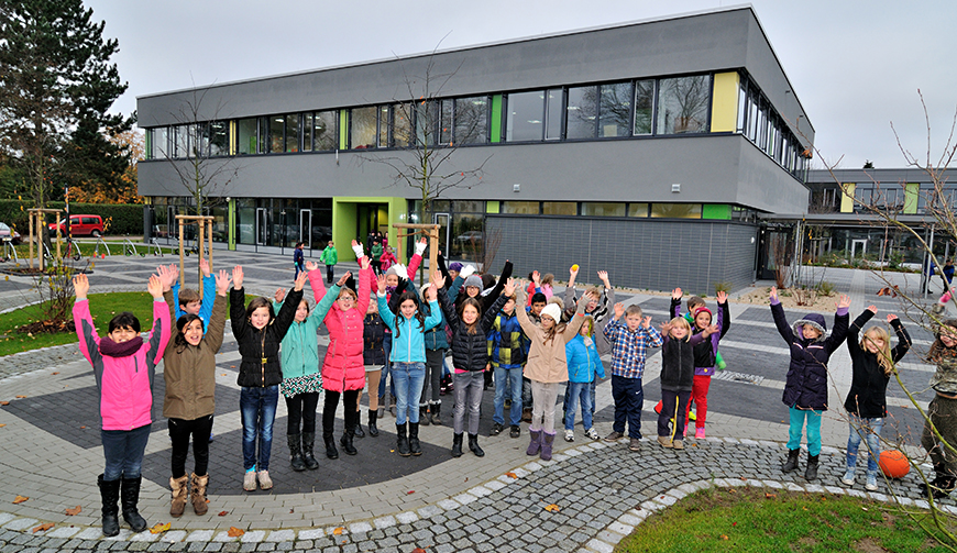 eine Gruppe Kinder, die jubelnd auf einem gepflasterten Hof vor einem grauen Gebäude mit grünen und gelben Verzierungen steht.