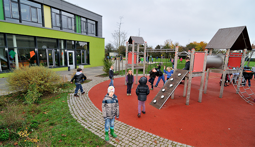 rechts im Bild spielende Kinder auf einem Spielplatz mit Klettergerüst, links im Bild ein grau-grünes Gebäude.
