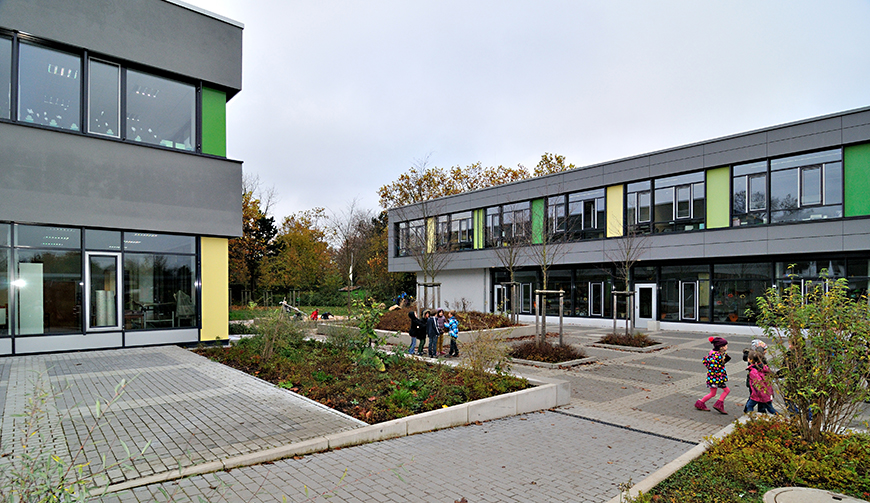 einen gepflasterten Innenhof zwischen zwei grau-grünen Gebäuden, auf dem etwas verdeckt von zwei bepflanzten Beeten einige Kinder stehen.