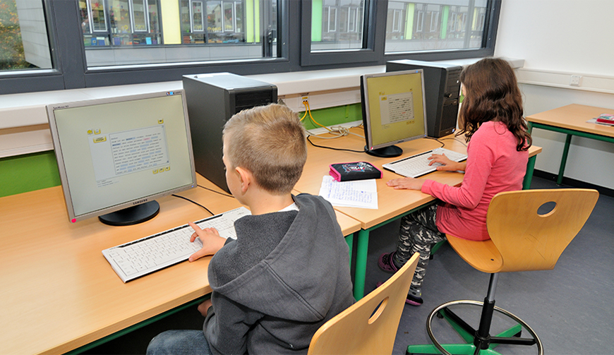 zwei kleine Schreibtische mit Computern, die vor einer Fensterfront stehen. An den Schreibtischen sitzen ein Junge und ein Mädchen und bearbeiten auf dem Bildschirm angezeigte Texte.