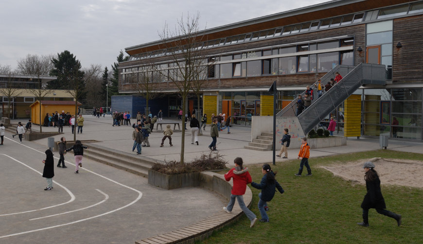 einen großen Hof auf mehreren Ebenen mit einer Rasen- und einer Sandfläche; überall spielen Kinder und im Hintergrund steht ein großes Gebäude mit einer Außentreppe