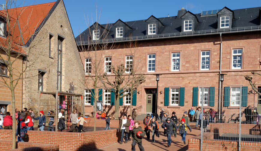 den Blick über einen rot gepflasterten Schulhof, auf dem Kinder spielen, auf zwei Backstein-Häuser