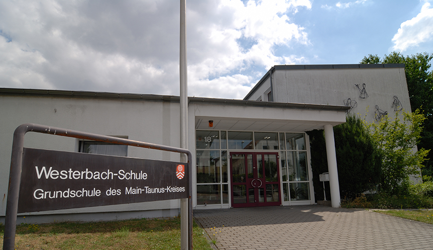 den Blick auf den verglasten Eingang eines Gebäude; links daneben ein großes Schild mit dem Text Westerbach-Schle - Grundschule des Main-Taunus-Kreises