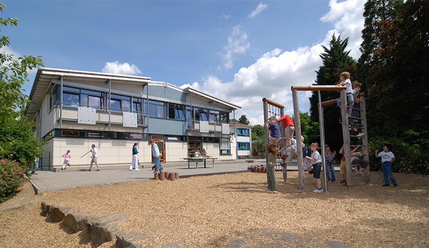 einen Spielplatz mit Klettergerüst, auf dem Kinder spielen; im Hintergrund ein großes Gebäude