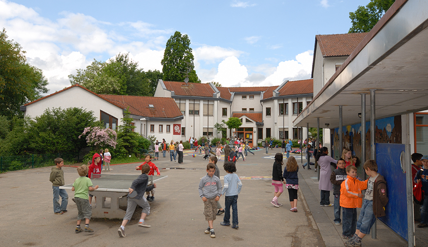 Blick auf einen großen Hof mit Tischtennisplatte, in dem viele Kinder spielen; im Hintergrund Häuser