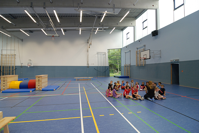 eine große Turnhalle mit blauem Boden, in der eine Gruppe Kinder im Kreis auf dem Boden sitzt