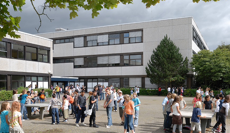 viele Schüler in einem Pausenhof mit Tischtennisplatten, im Hintergrund ein Gebäude