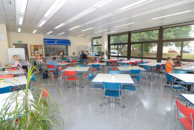 Blick in einen großen Raum mit vielen kleinen Tischen, um die blaue und rote Stühle stehen; im Hintergrund befinden sich einige Schüler