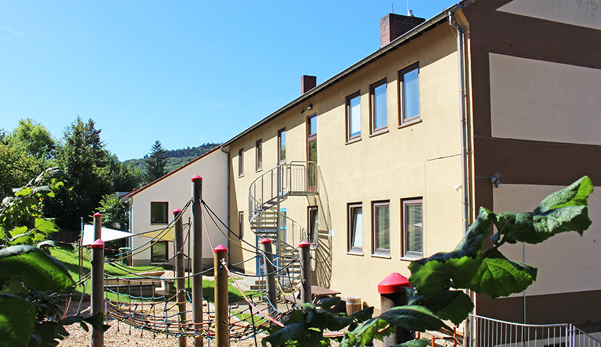 ein gelb gestrichenes Haus mit Wendeltreppe außen, davor ein Garten mit einem Klettergerüst