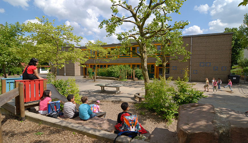 Blick in einen Schulhof mit Bäumen und Hecken, im Vordergrund einige Kinder an einem Klettergerüst, im Hintergrund ein Gebäude