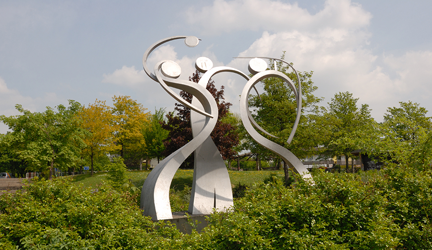 eine große Skulptur aus Edelstahl inmitten einiger Hecken