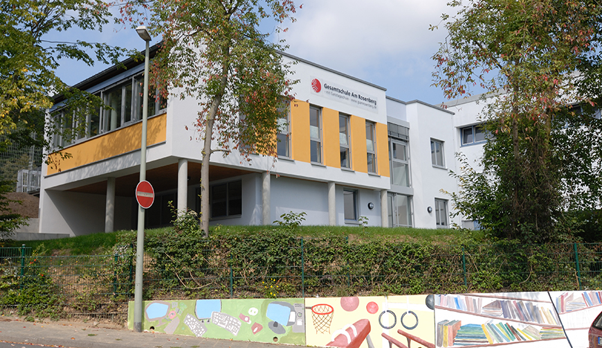 Blick auf eine bunt bemalte Mauer, dahinter eine Hecke und dann ein weiß-gelb gestrichenes Gebäude