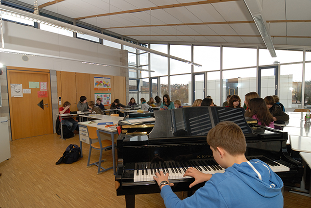 im Vordergrund einen Schüler an einem Klavier und im Hintergrund einige weitere Schüler an Tischen