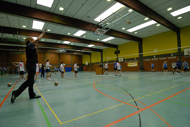 den Blick in eine Turnhalle; im Vordergrund gibt ein Trainer Anweisungen, im Hintergrund bewegen sich Sportler