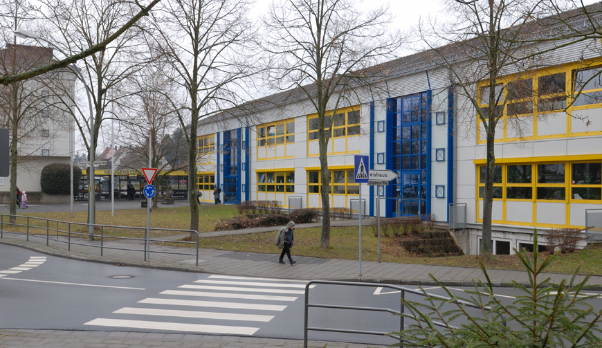 im Vordergrund eine Straße mit Zebrastreifen, im Hintergrund Bäume und Wiese vor einem Gebäude