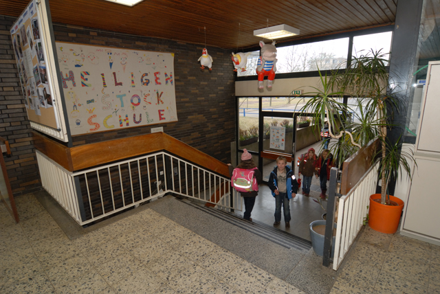 Blick aus dem ersten Stock eine Treppe hinunter in eine Eingangshalle, wo sich einige Schüler befinden