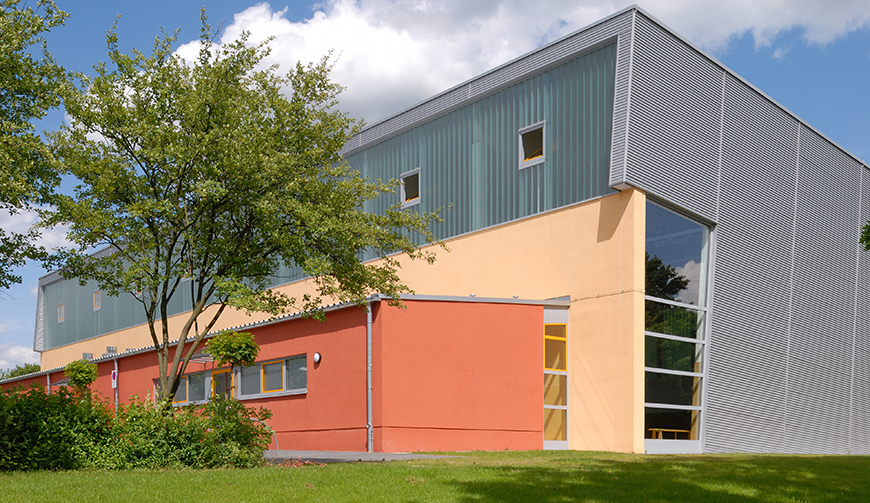 Blick über eine Wiese auf ein großes graues Gebäude mit einem orange-roten Anbau
