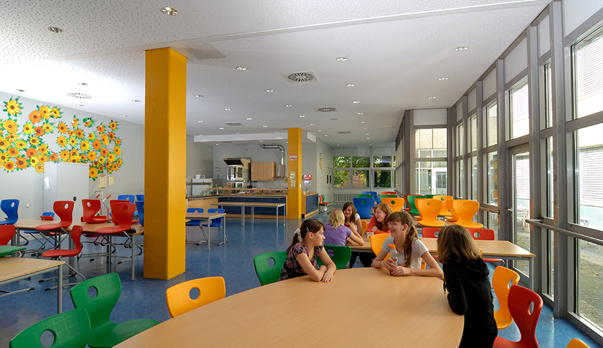 Blick in einen großen Raum mit Holztischen und bunten Stühlen, an denen einige Kinder sitzen