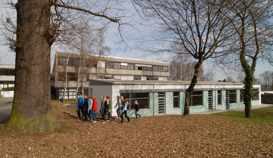 den Blick über eine Wiese mit Bäumen, auf der eine Gruppe Kinder läuft; im Hintergrund einige Gebäude