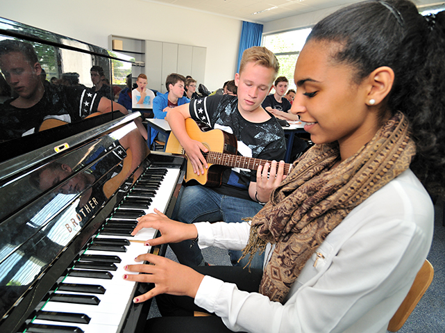 im Vordergrund eine Jugendliche an einem Klavier, im Hintergrund ein Schüler mit einer Gitarre; im Raum sitzen weitere Schüler