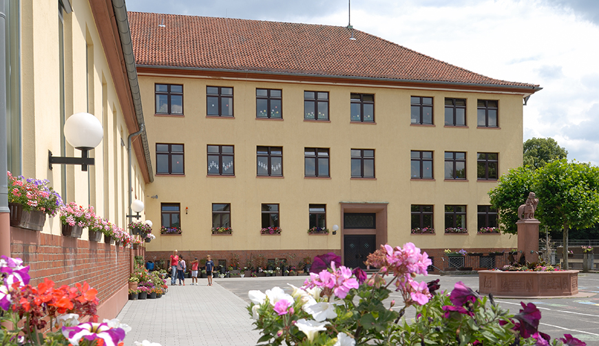 Blick über einige Blumen auf einen Hof mit Brunnen und ein großes, gelb gestrichenes Haus