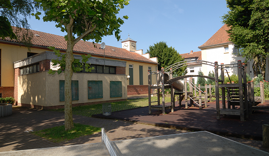 den Blick auf einen Innenhof mit Spielgeräten; im Vordergrund eine Tischtennisplatte, dahinter ein Klettergerüst und im Hintergrund ein Gebäude