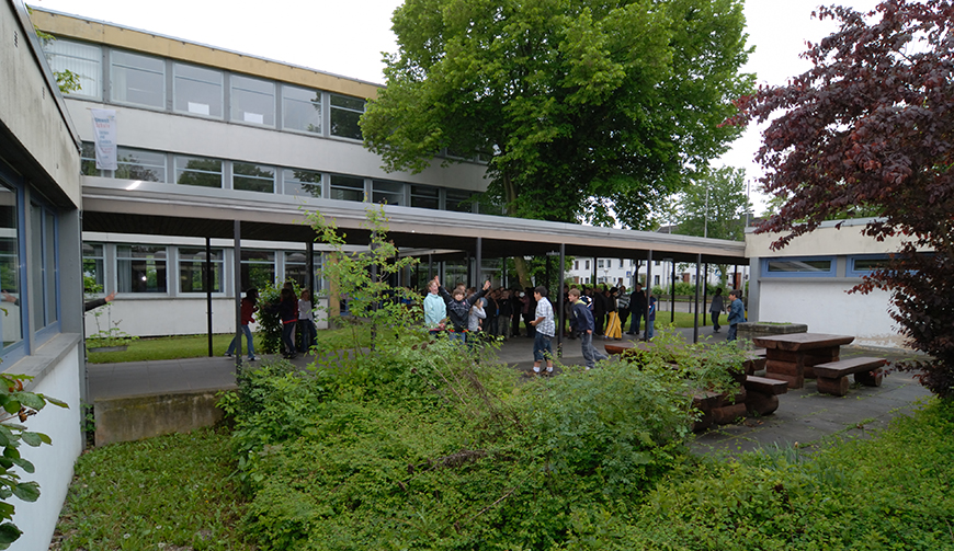 Blick auf einen mit Hecken bepflanzten Hof, in dem einige Tische und Bänke stehen; im Hintergrund einige Schüler vor einem Gebäude
