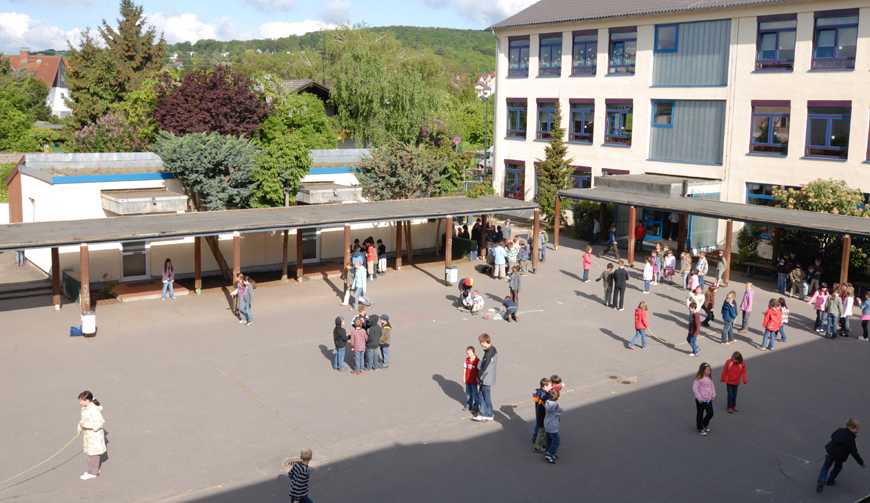 Blick von oben auf einen Hof, in dem Kindergruppen stehen und spielen, im Hintergrund Gebäude und Bäume