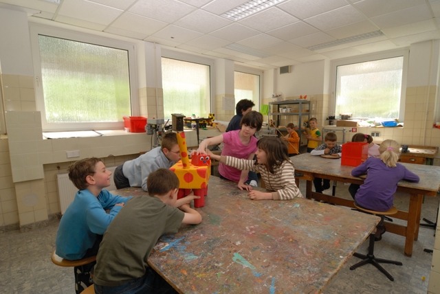 einen Raum mit vielen Fenstern und einigen bunt beschmierten Holztischen, an denen Kinder arbeiten