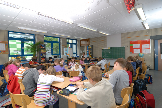 einen Klassenraum mit Holztischen und -stühlen, an denen viele Kinder sitzen; im Hintergrund eine Fensterfront und eine Tafel
