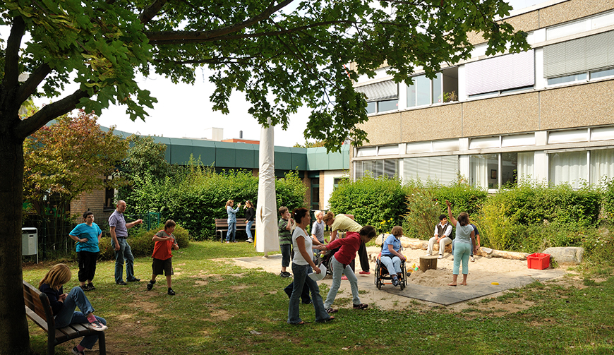 eine Wiese und einen Sandkasten, wo einige Kinder spielen; links ragt ein Baum ins Bild, im Hintergrund steht ein Gebäude