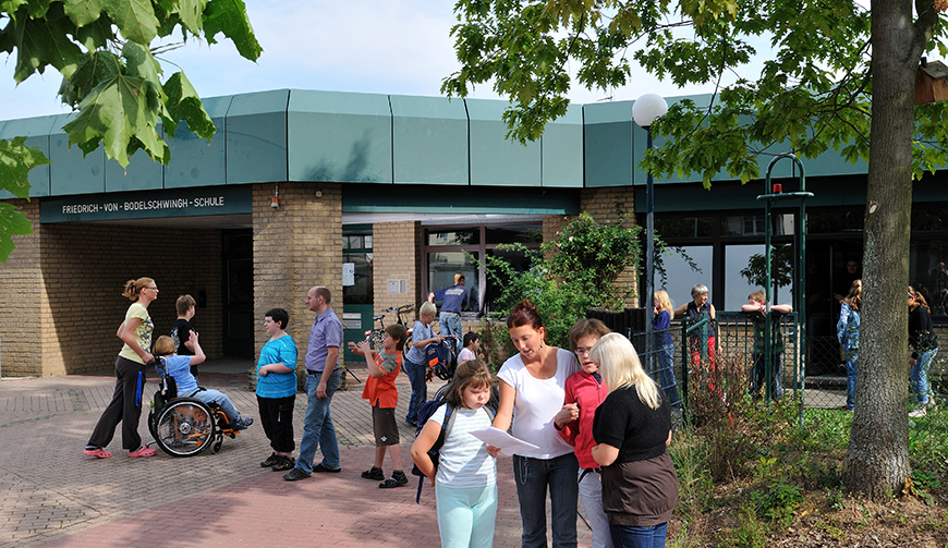 Kinder und Erwachsene in einem von Bäumen gesäumten Hof vor einem niedrigen Gebäude