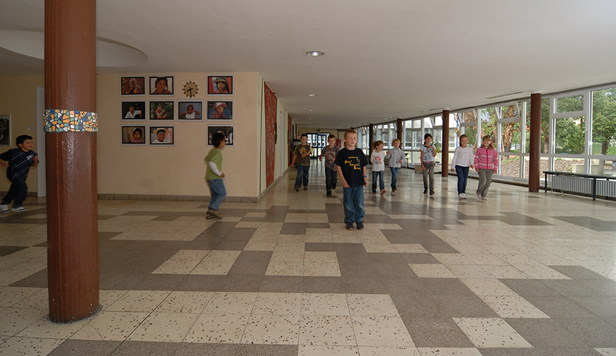 eine Eingangshalle mit vielen Fenstern und einer Säule, in der einige Schüler auf die Kamera zulaufen
