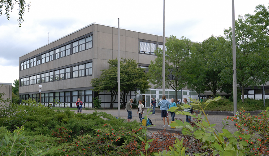 Blick über ein grün bepflanztes Beet und eine Gruppe Schüler hinweg auf ein graues Gebäude