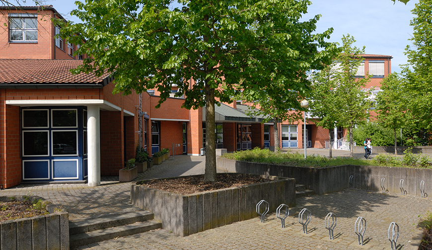 einige Fahrradständer in einem Hof; hinter einem Baum ein großes rotes Gebäude