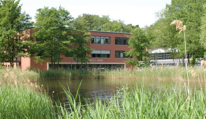 den Blick über einige Gräser auf einen großen Teich; im Hintergrund ein großes rotes Gebäude