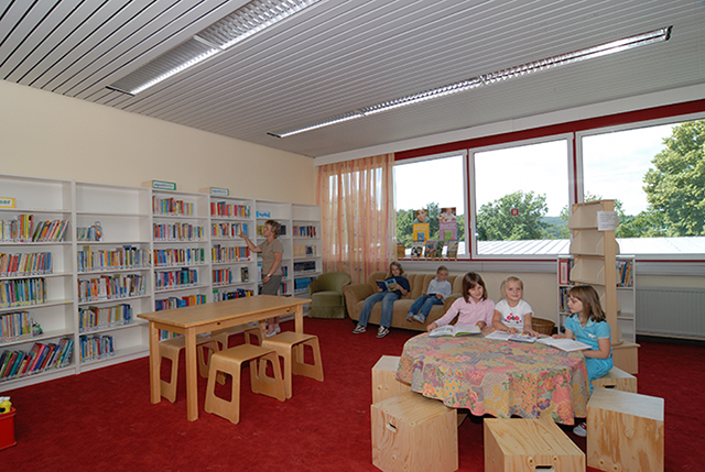 den Blick in einen Raum mit Holztischen und -hockern, an denen einige Kinder sitzen; im Hintergrund Bücherregale