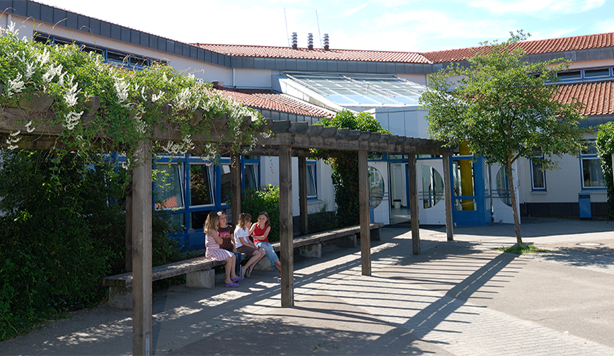 den Blick über einen Hof mit einer berachsenen Holz-Pergola, unter der Jugendliche auf Bänken sitzen